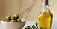 olivas picual: Tazón blanco con aceitunas verdes junto a una botella de aceite de oliva y ramas de olivo.