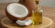 Beneficios del aceite de coco en el ombligo: Mitades de coco y frasco con aceite sobre tabla de madera, iluminación suave y fondo neutro.